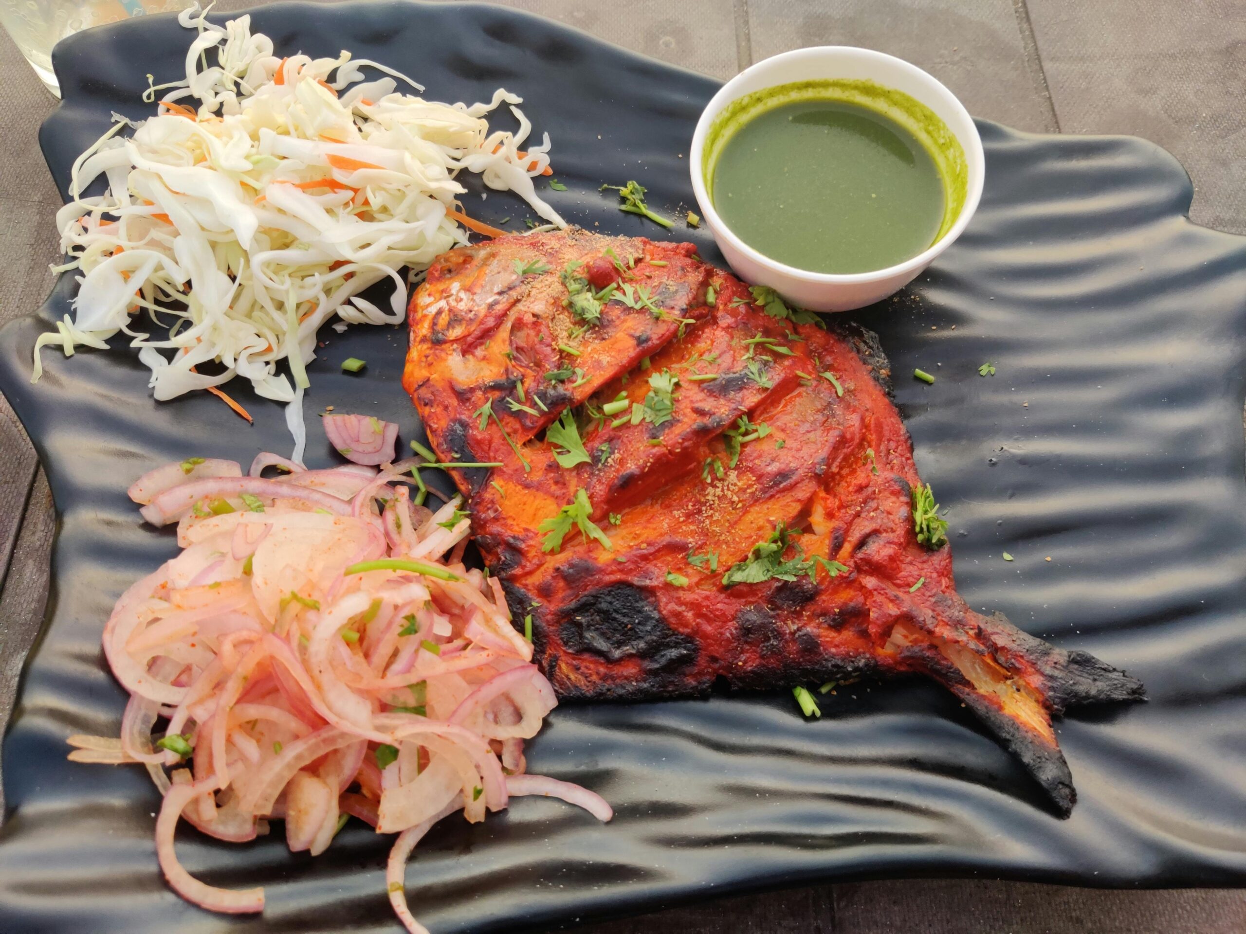 Grilled tandoori chicken leg on a black plate with green chutney, shredded cabbage and onion salad on the side.