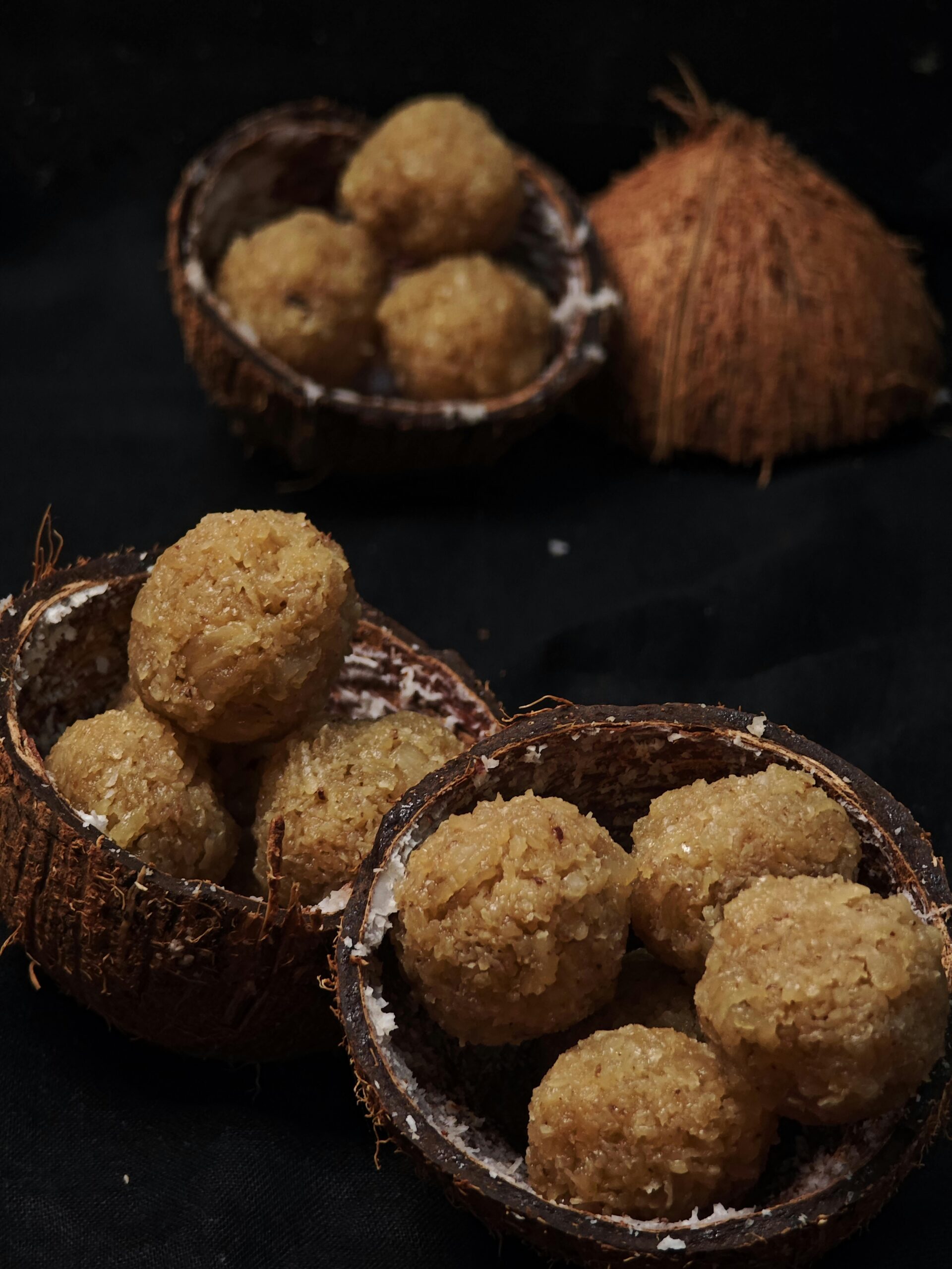 Two halved coconuts used as bowls holding beige crumbly coconut candy balls on a dark surface.